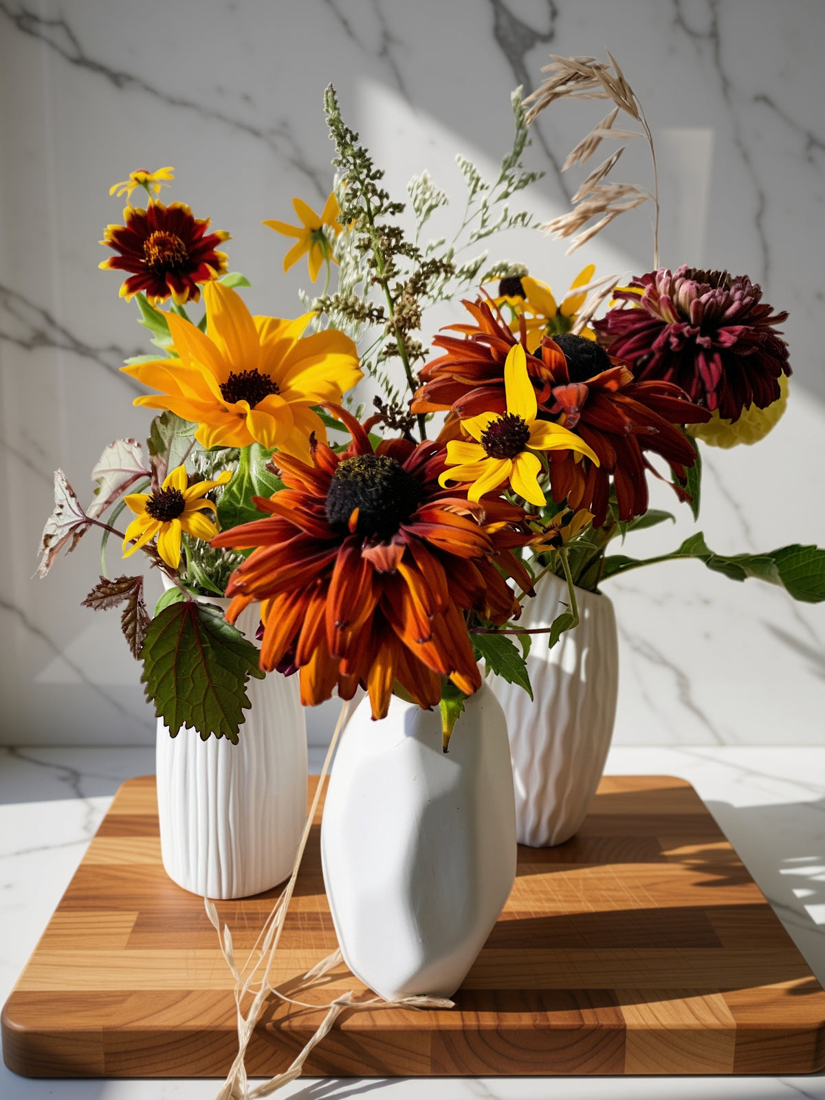 A floral arrangement of yellow, orange, and burgundy flowers in three white ribbed ceramic vases placed on a wooden cutting board against a white marble wall.