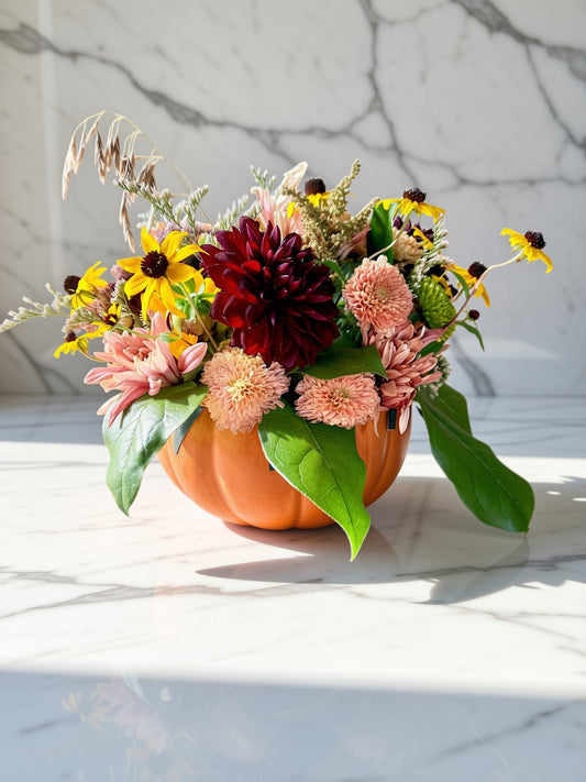 An autumn floral arrangement featuring yellow, burgundy, and pink flowers with green leaves, styled inside a carved orange pumpkin on a white marble surface.